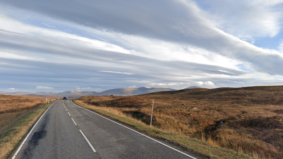 A general view of the A82 road in Argyll and Bute. The road goes through the middle of the picture. On either side are brown and green grasses. Hills slightly lit up by the sun can be seen in the background. The sky is blue and streaked with white cloud.