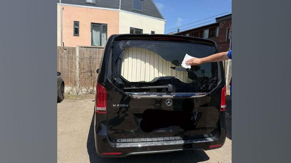 A back view of a black Mercedes vehicle in a car park. Its rear window has been smashed leaving a hole across two thirds of it. A man's arm can be seen to the left, reaching across with a cloth to wipe the remains of the black glass.