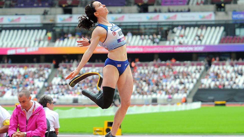 Stef Reid, wearing a blade and a trainer while jumping. There is a stadium behind her and an umpire dressed in a pink top.