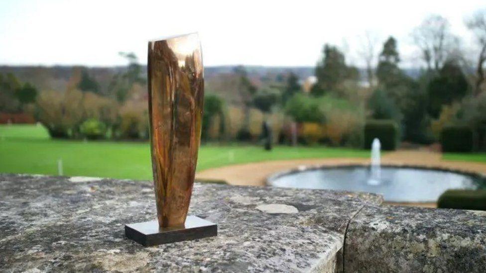 A plain polished bronze sculpture on a stand. It appears to be the size of a forearm and has been placed on a stone-capped wall in the open air. In the background is a lawn with a round pond and fountain.