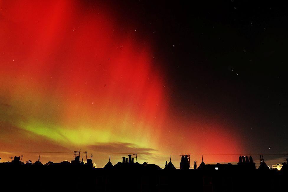 The orange and green glow of the Northern Lights over silhouetted rooftops.