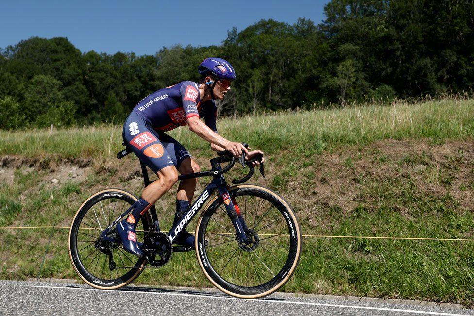 Cyclist Oscar Onley viewed side on on a Lapierre bike in blue and orange kit against a green countryside background