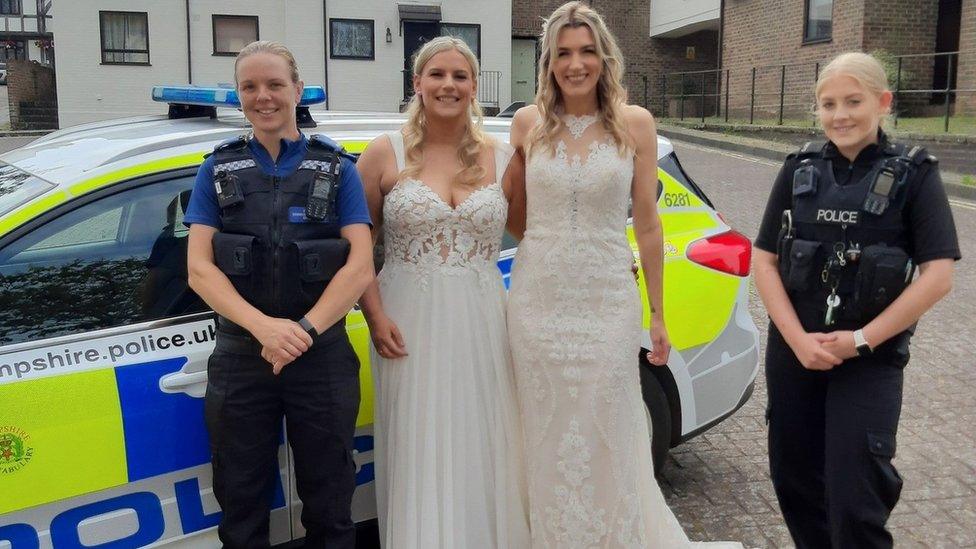 Two women wearing wedding dresses standing between two women police officers in front of a police car