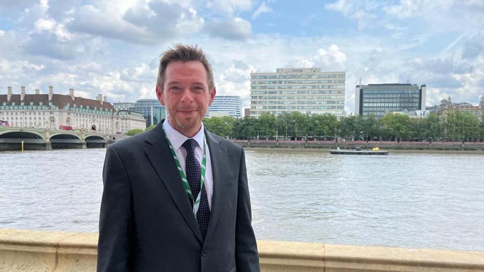 A man with short hair and wearing a dark suit, pale pink shirt, black tie and green lanyard around his neck, stands next to a wall with a river, bridge and buildings in the background.