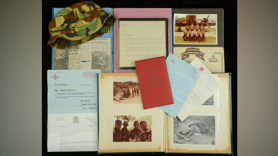 Army memorabilia belonging to Baz Grayling lying on a black backdrop. On the top row is a hat in camouflage on top of a yellowing press cutting, plus a photo and letter with Parachute Regiment insignia. On the bottom row are more letters and a photo album, open to show photos, plus more letters lying on top of the album.