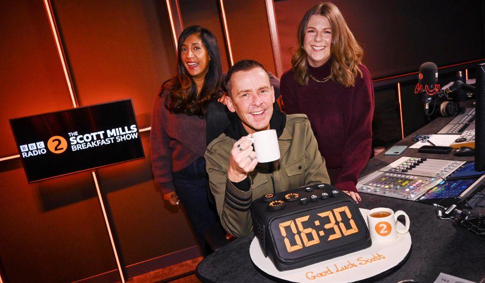 Scott Mills pictured in the Radio 2 studio with Tina Daheley and Ellie Brennan, and a cake in the shape of an alarm clock