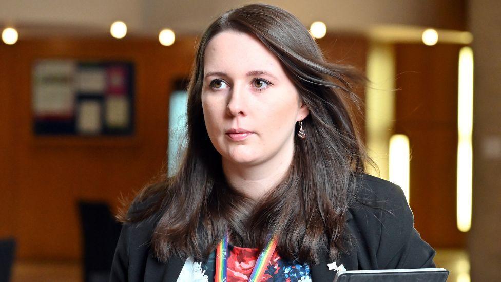 Emma Roddick, with dark hair and wearing a dark suit, walks in a corridor in the Scottish Parliament 