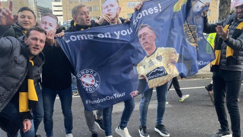 A group of maidstone fans holding a flag showing the face of Maidstone midfielder Sam Corne