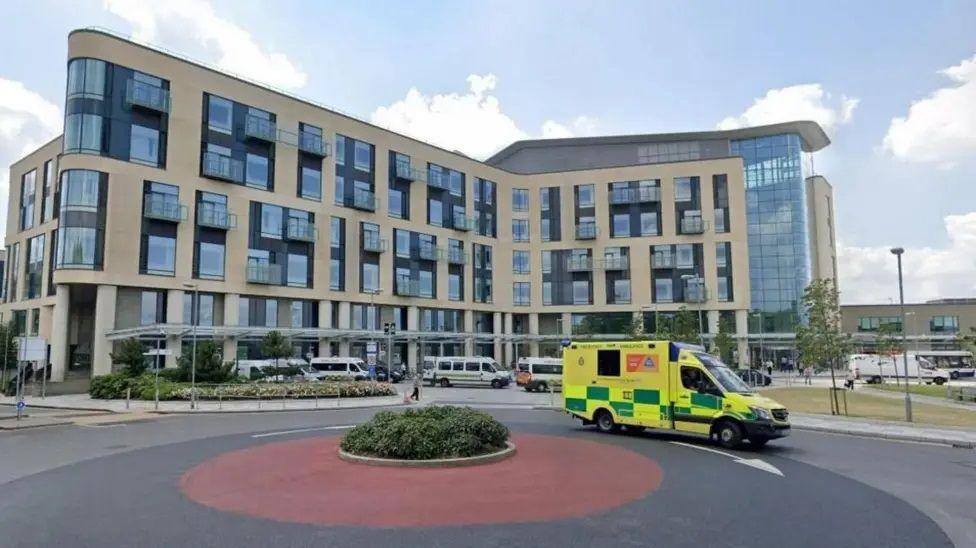 The outside of Southmead Hospital. The building is modern and has lots of large windows and a tall section made entirely of glass. There are a number of hospital transport vans parked outside. In the foreground there is a red mini roundabout with a small hedge in the centre. A bright yellow and green ambulance is seen driving around it. 