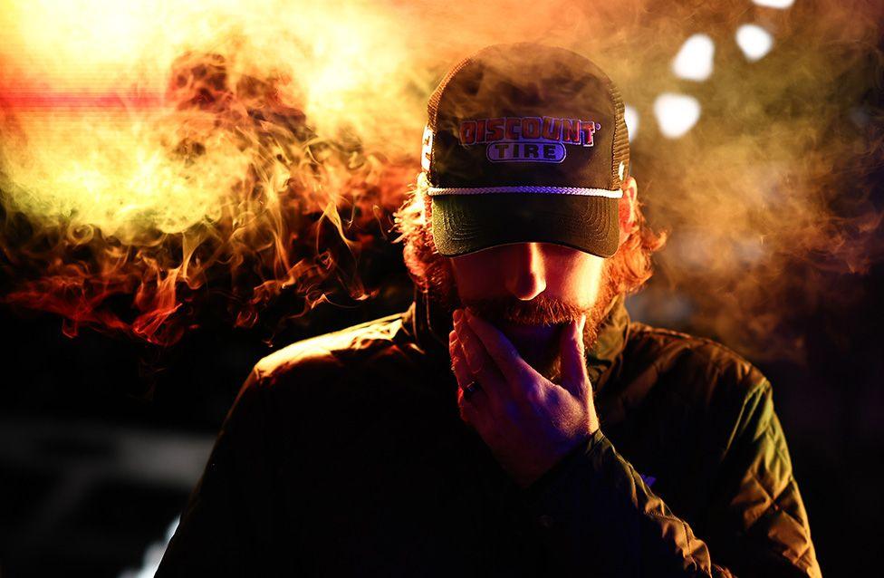 A male driver wearing a dark jacket and a cap with a logo, surrounded by swirling orange and yellow smoke against a dark background with bright light spots