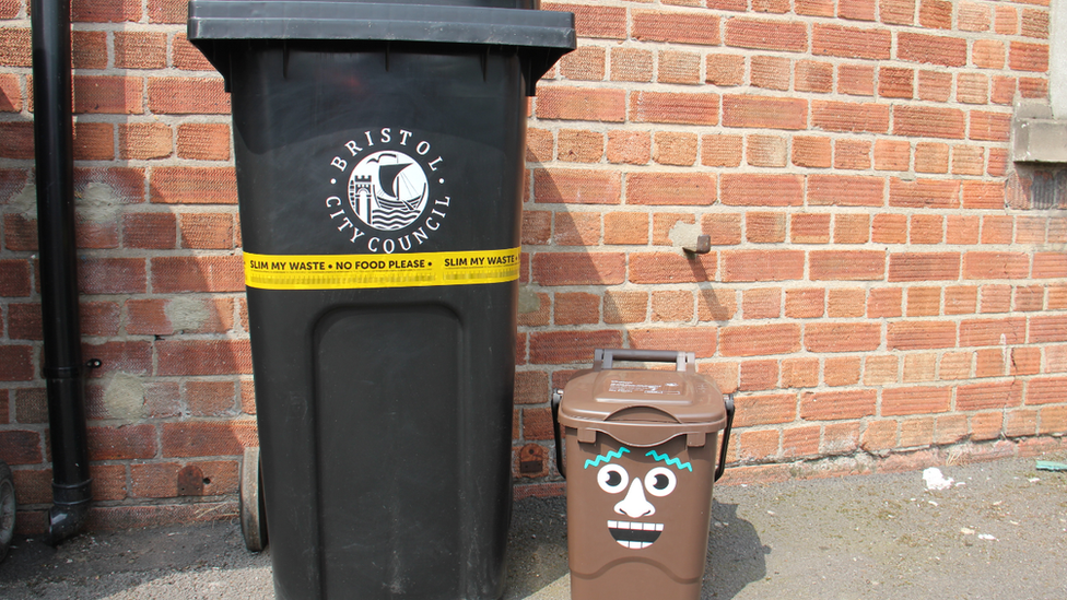 A black bin with the Bristol City Council logo on it. Wrapped around the bin is a yellow plastic ribbon that reads 'Slim my waste, No food waste'. Next to it is a small brown food waste bin with cartoon face stickers on it.