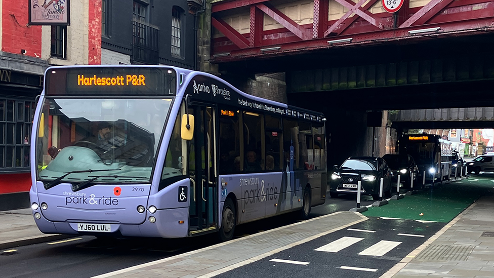 A single lane road with a bus stop in the middle. A Harlescott Park & Ride bus is waiting there, with a line of traffic behind it. A 1.1m-wide pavement "island" is next to the bus. To the right of that is a cycle lane, which is coloured green, apart from a section with a small, two-striped section of zebra crossing