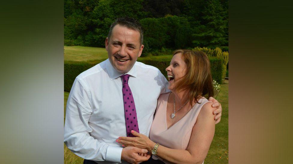 John Hunt with his left arm around Carol Hunt shoulder and her left hand touching his midriff. He is smiling at the camera; she is laughing while looking at him. She is wearing a V-neck peach coloured top with a necklace; he is wearing a white shirt with a mauve tied with black spots on it