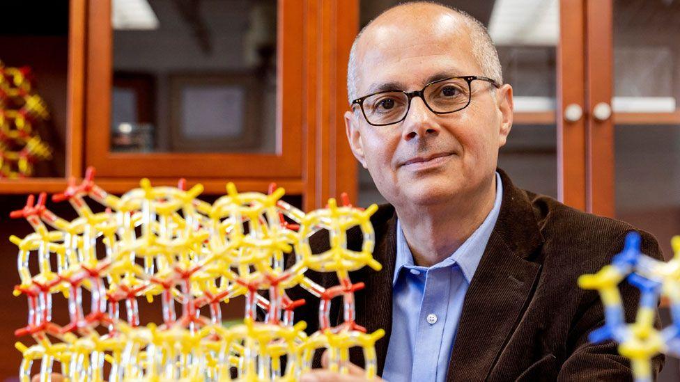UC Berkeley chemistry professor Omar Yaghi, wearing a blue shirt, brown jacket and glasses, poses in his lab at the University of California in Berkeley US. Next to him is a model showing metal-organic frameworks made of yellow and red tubes and nodes.