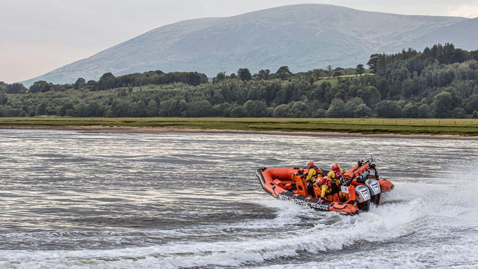 A crew on the Nith Inshore Rescue boat make a sharp turn on the water with a tree-lined shore and hill in the background