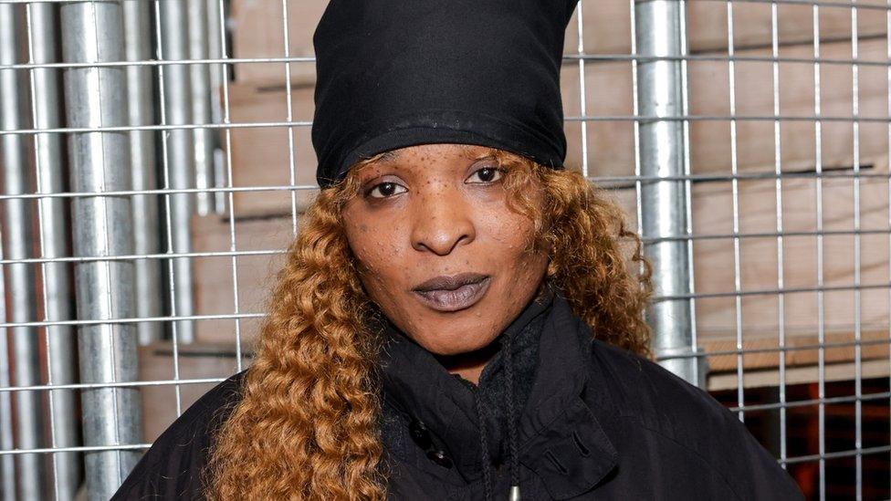 Torishéju Dumi. Torishéju is a 31-year-old black woman with brown eyes, freckled skin and long, curly ginger hair. She wears a black hat and black puffer coat and is pictured in front of a metal grate, behind which some stacked plywood boxes can be seen.