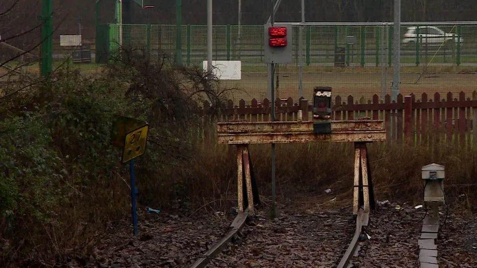 Barriers and a fence at the end of a railway line. In the background, a car is driven across a road.