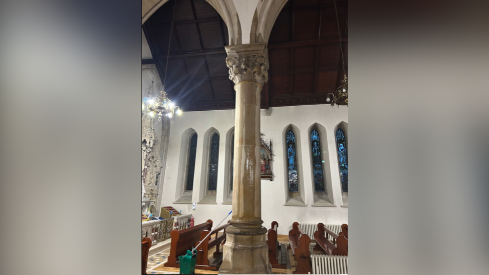 Water trickling down internal church pillar. A water bucket and mop is next to it. We can see pews just beyond the pillar, and then a white wall in which several stained glass windows are set. 