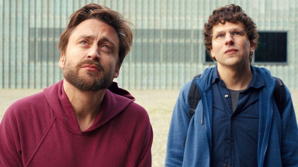 Kieran Culkin and Jesse Eisenberg in A Real Pain - Culkin is wearing a maroon hoodie and Eisenberg is wearing a blue one.