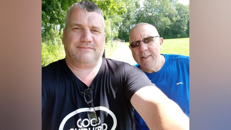 Ade Doughty stands in a black tshirt that reads "good shepherd". He has grey hair. he is next to a man in a blue t-shirt and black glasses with a bald head. Behind them is a path, green grass and trees