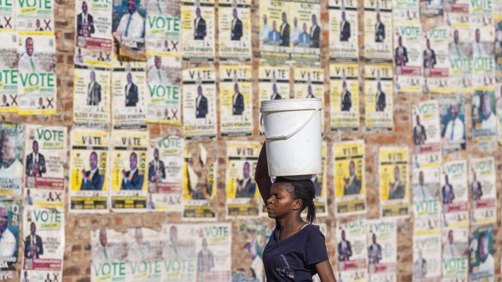 A woman carrying a container filled with water passes in front of a wall with campaign posters of participating candidates in the country's 2023 general elections in Harare, Zimbabwe - 18 August 2023