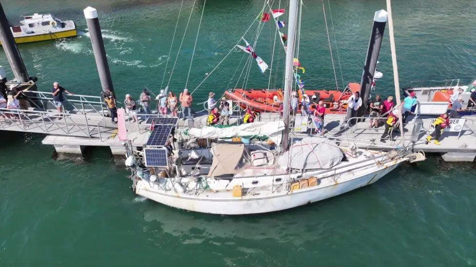 This picture shows a sailing boat moored at a dock with solar panels on the deck of the sailboat. Various pieces of equipment are visible, such as lifebuoys and ropes. Colourful flags are hanging from the mast and there are several people on the dock.