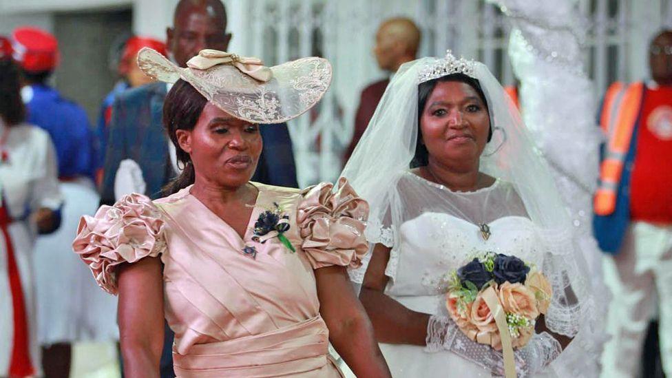 A woman (L) dressed in a beige silky dress with ruched short sleeves and a hat stands beside a bride in a white wedding dress, white lace gloves who smiles as she looks at the camera, holding a bouquet of flowers, including orange roses.