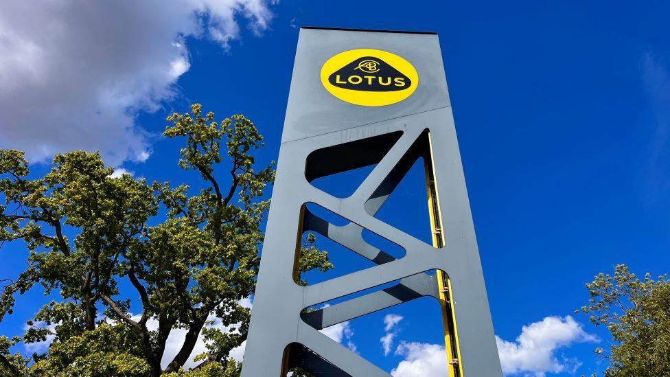 A tall grey sign outside the Lotus headquarters in Hethel, near Wymondham, in Norfolk. The sign has a Lotus logon which is yellow with a black curved triangle inside which says LOTUS in yellow writing. There are green trees surrounding the sign and the sky is blue as the photograph has been taken during the day.