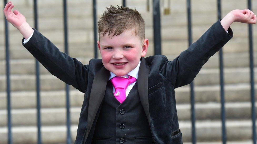 Dáithí MacGabhann smiles with his arms outstretched. He is wearing a black three-piece suit and a pink tie.