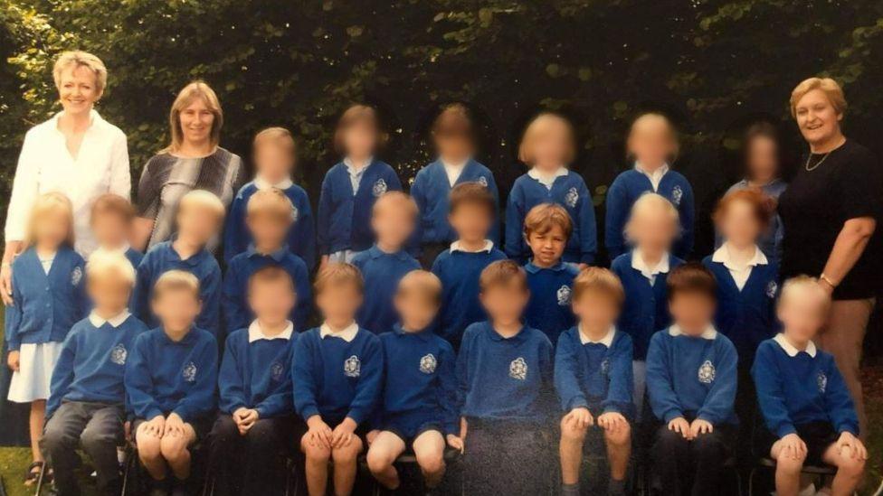 A class picture taken when Lando Norris was a pupil at the primary school. There are three rows of children wearing blue sweatshirts with the school logo on it. They are standing outside with trees in the background. Three female teachers stand at the back of the children smiling at the camera. All of the children's faces have been blurred except for Lando Norris.