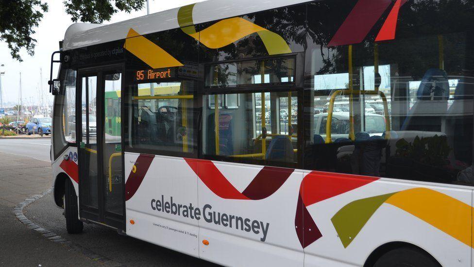 A white bus with red, yellow and green ribbon-style livery is driven from the right with a small section of road seen on the left. celebrateGuernsey is written on the livery. 