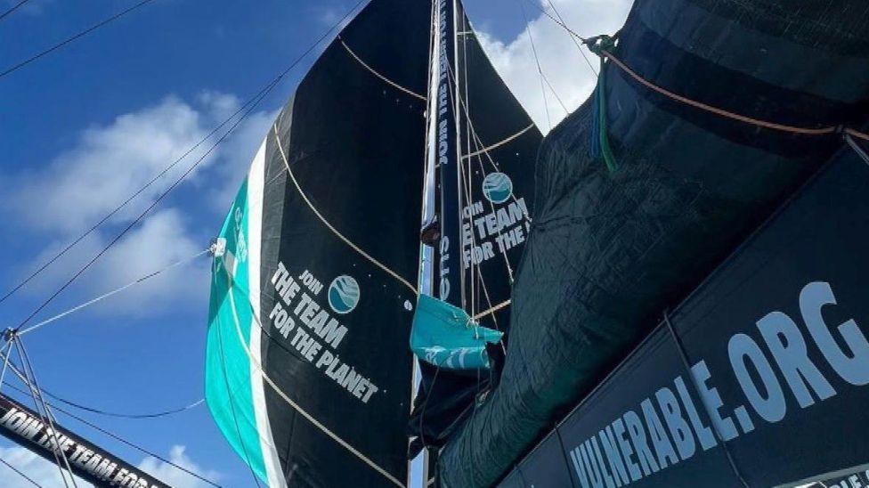 Two jibs on either side of the mast with a mainsail folded up and a number of written logos with blue sky above.