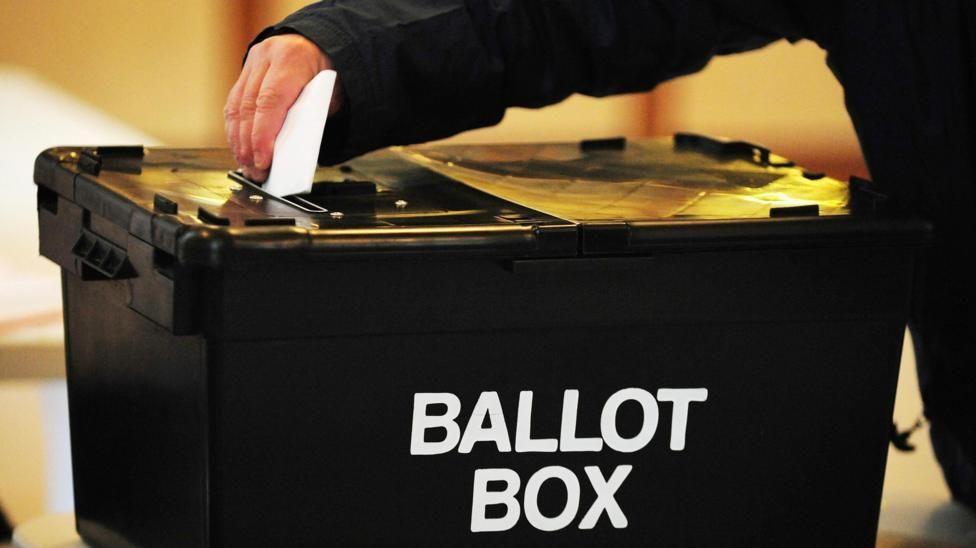 A man's hand holding a white piece of paper reaches across a black box, marked "Ballot Box" in white lettering, with a slot on the lid.