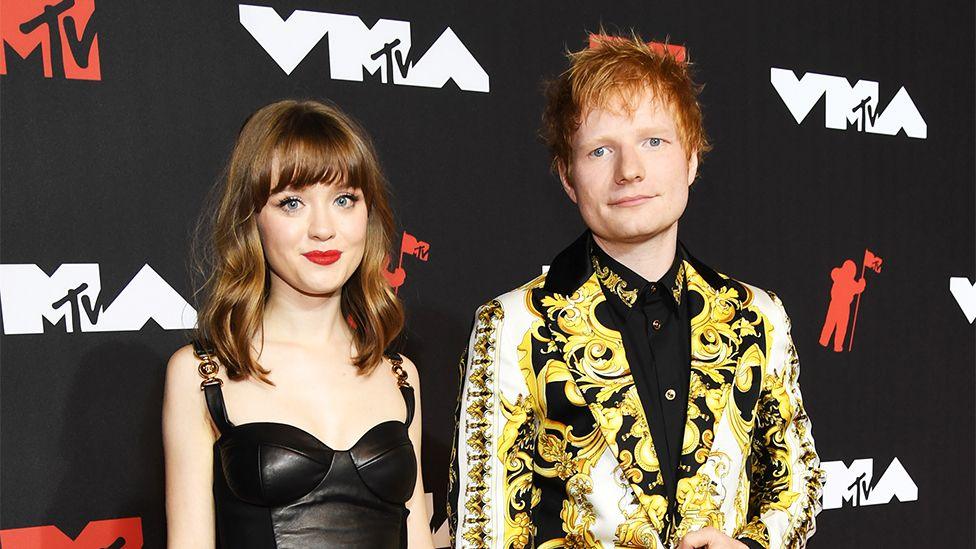 Maisie Peters and Ed Sheeran on the red carpet at an MTV event. A young Maisie Peters has brown hair and wears black ankle boots with heels and a black leather dress with buckle straps. Ed Sheeran wears a yellow paisley patterned blazer with black trousers and shoes and a black shirt underneath