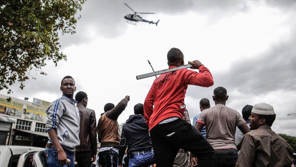 Helicopter flies low as group of men, some armed, stand in a crowd