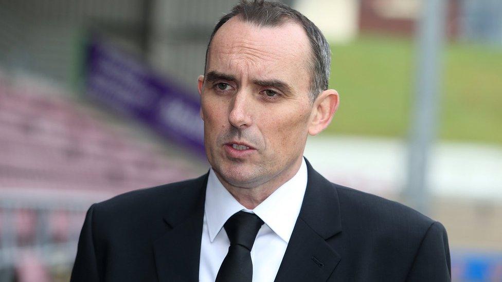 Man with short dark hair wearing a black suit and black tie