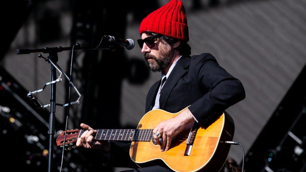 Gruff Rhys on stage in a red bobble hat and suit, playing a guitar