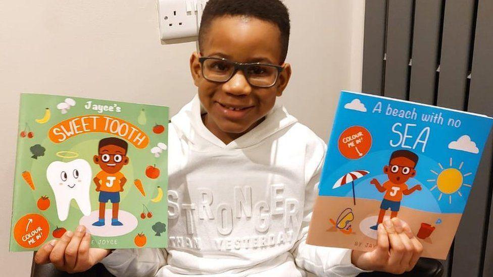 Jayce, wearing glasses and a white top, smiles at the camera while holding a a book in each hand. One is Jayce's Sweet Tooth and the other is A Beach With No Sea.