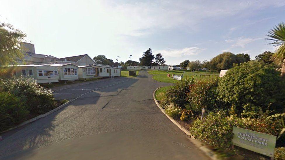 Green holiday park sign in shrubs on right with road leading up to lines of cream-coloured caravans