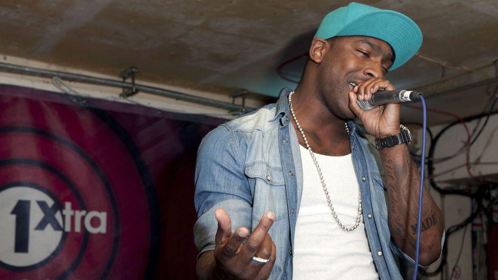 A young Skepta, wearing an open denim shirt over a white t-shirt, rapping into a microphone in the BBC Radio one extra studios.