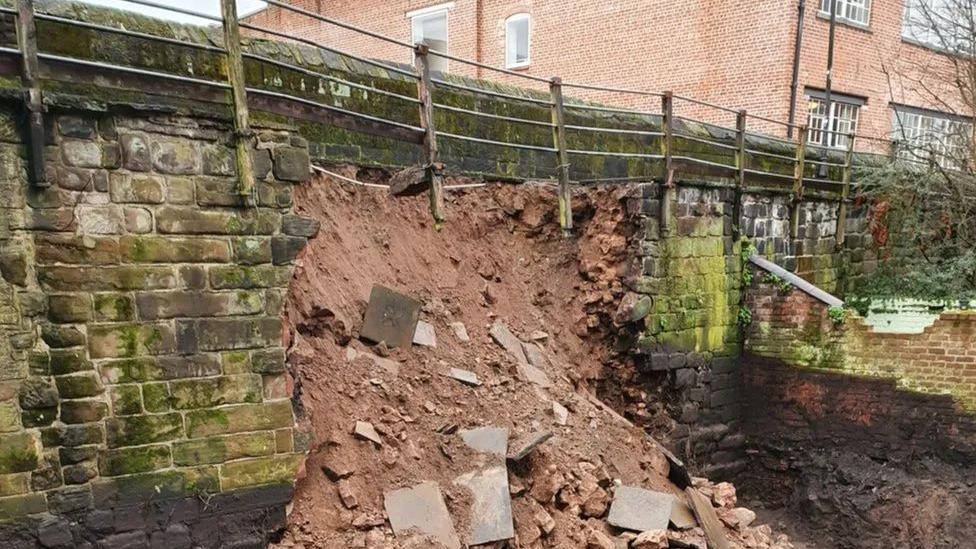 Rubble and slabs of orangey brown concrete have broken away from a walkway of bricks. Silver railings guard the edge to the walkway.