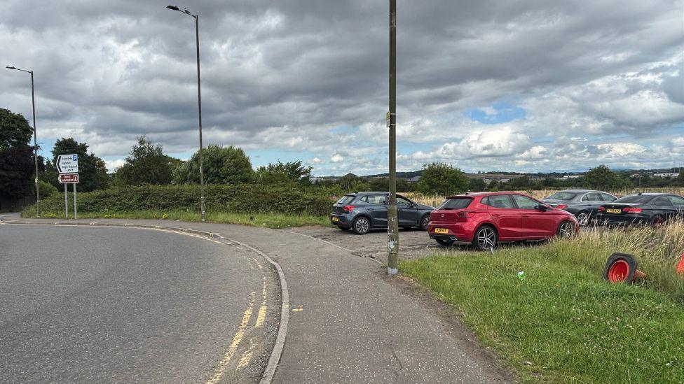 A layby near a roundabout at Edinburgh Airport. There are four cars. The car closest to the camera is red. The cars further away are dark coloured, either black or grey. 