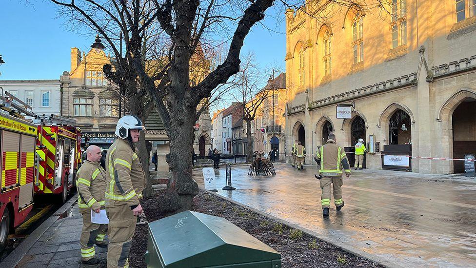 About half a dozen firefighters are in the vicinity of the damaged building during daylight. There is red and white tape cordoning off the area. Two fire engines are parked at the scene.