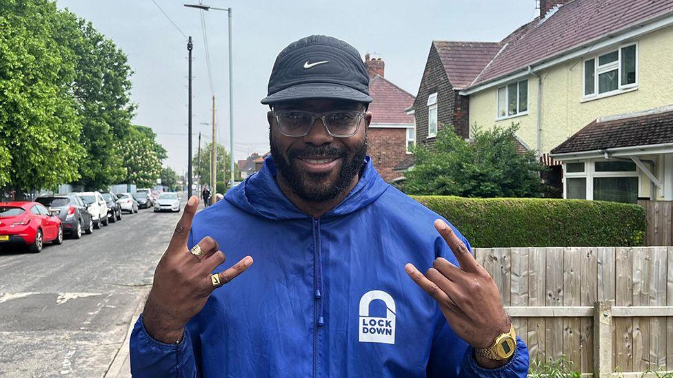 Chiedu Oraka standing in a Hull street he has a beard and wears glasses, a baseball hat and blue-hooded jacket