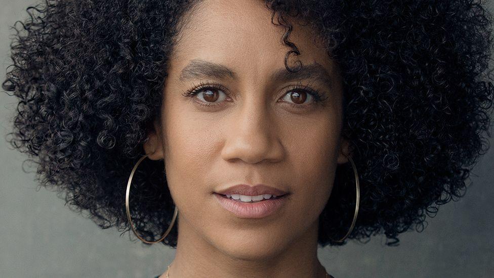 A professional headshot of a woman with dark, curly hair. She's wearing large hoop earrings and her expression is neutral. 