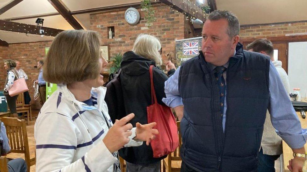 Woman in white top with arms outstretched talks to minister in blue gilet leaning on a chair.