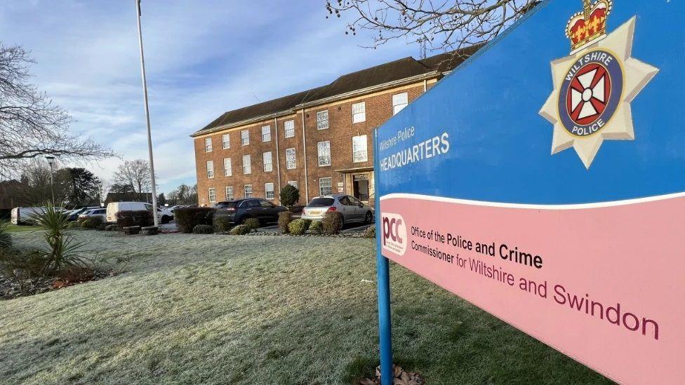 The outside of Wiltshire Police's headquarters. A blue and pink sign indicating it is the headquarters is outside the building in the foreground, while the building itself is in the background. There are a number of cars and vans in front of the building.