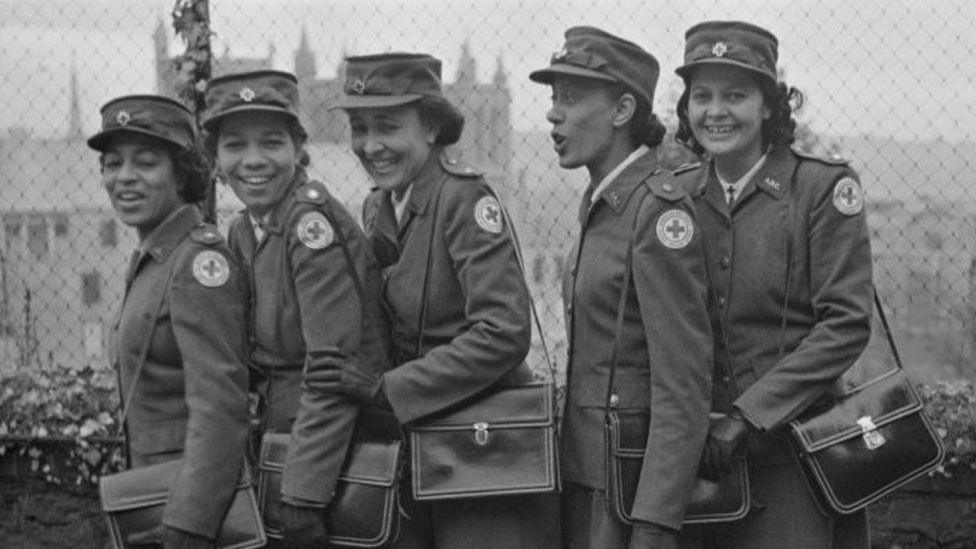 Five American Red Cross servicewomen. They are in uniform, with peaked caps, and leather bags over their shoulders, standing sideways on. Behind them can be glimpsed buildings. Left to right: Sydney Taylor Brown, of Pittsburgh, Pennsylvania, Henrine Ward, of Chicago, Illinois, Carol Jarett, of Denver, Colorado, Magnolia Latimer, of Atlanta, Georgia, and Gladys Edward Martin, of Topeka, Kansas. 