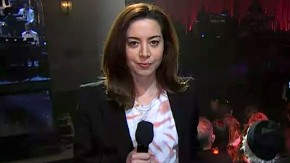 Wearing a tie dye t shirt and black blazer, Aubrey Plaza holds a microphine with the SNL stage behind her