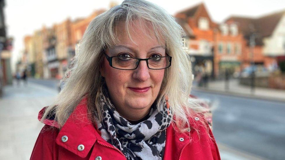 A blonde woman smiling. She is wearing glasses and a red coat